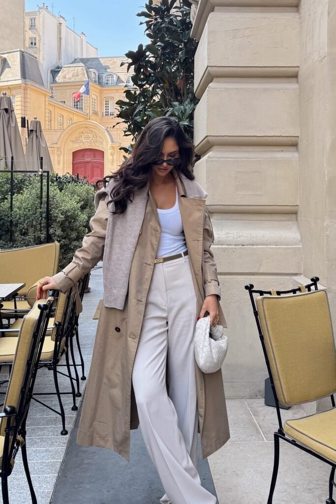 Beige Trench Coat + White Tank Top + Beige Wide-Leg Trousers + Beige Knit Sweater Draped Over Shoulders + White Woven Clutch Bag