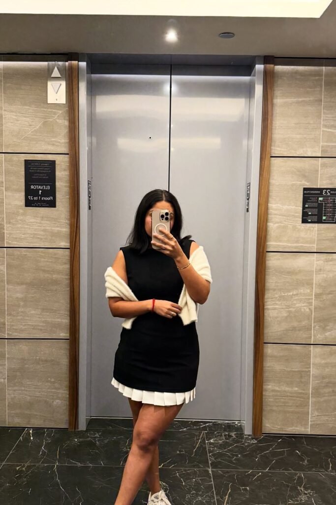 Black Pleated Hem Mini Dress + White Sweater Draped Over Shoulders + White Sneakers