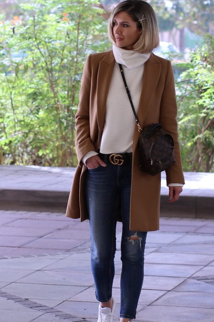 White Ribbed Turtleneck + Camel Long Coat + Blue Distressed Skinny Jeans + White Sneakers + Brown Monogram Mini Backpack