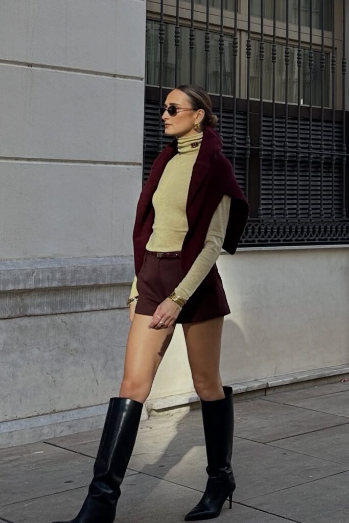 Light Yellow Turtleneck + Burgundy Sweater Draped Over Shoulders + Burgundy High-Waisted Shorts + Black Knee-High Boots + Gold Accessories + Sunglasses