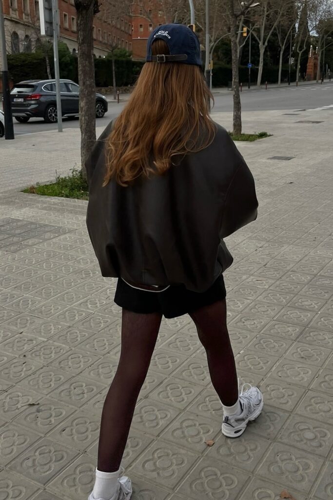 Oversized Dark Brown/Black Leather Jacket + Black Shorts + Sheer Black Tights + Navy Baseball Cap + Grey/White Trainers