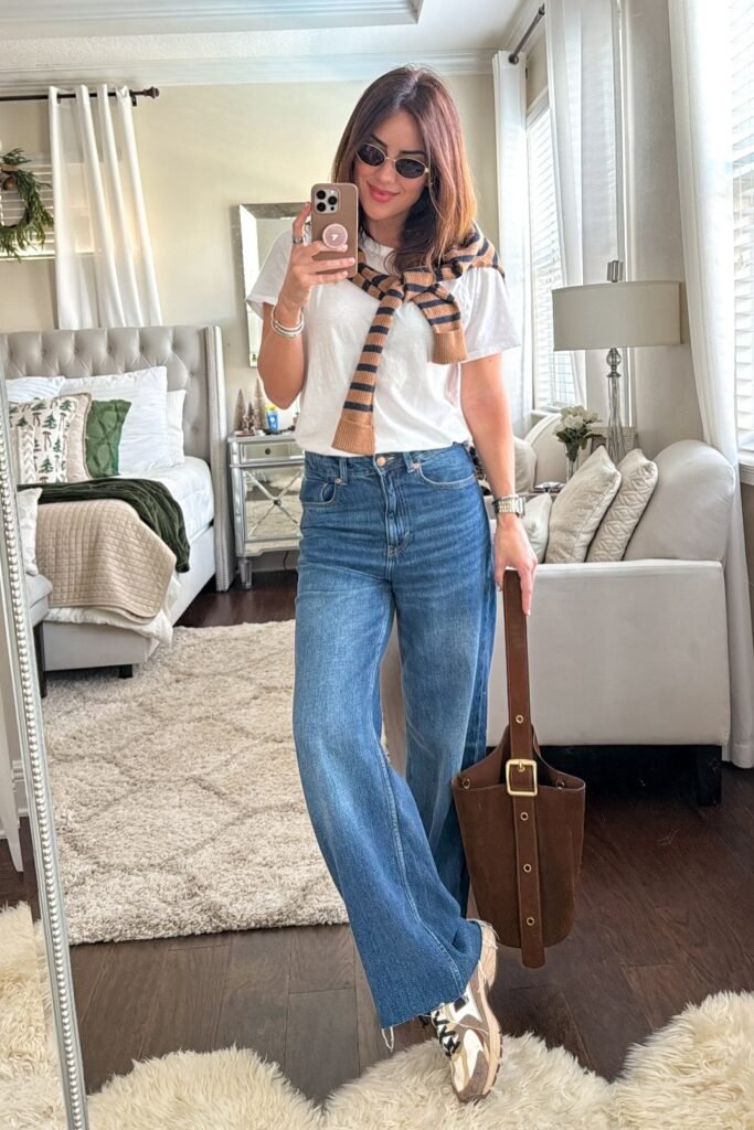 White Basic T-Shirt + Striped Sweater Over Shoulders + Mid-Wash Flared Jeans + Tan and White Trainers