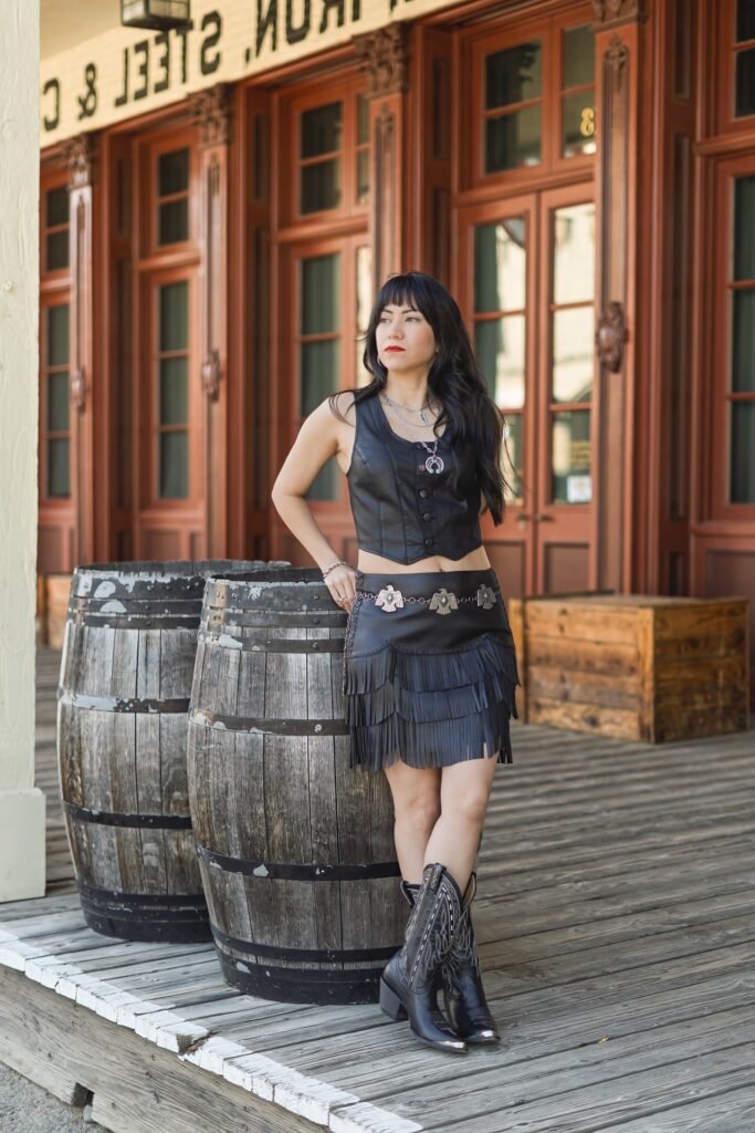Black Leather Button-Up Vest + Black Fringed Mini Skirt + Black Patterned Cowboy Boots + Silver Concho Belt