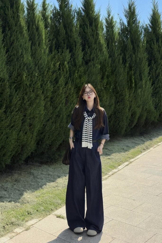 Dark Blue Denim Utility Jumpsuit + Striped Knit Sweater Slung Over Shoulders + Round Eyeglasses + Grey Slip On Shoes