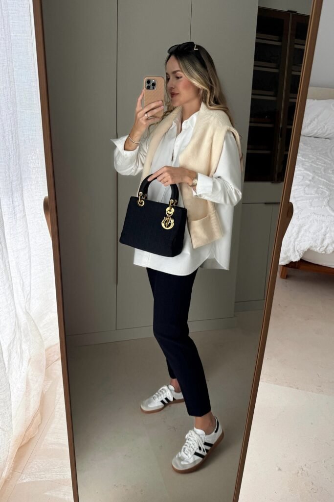 White Oversized Button Down Shirt + Cream Knit Sweater Slung Over Shoulders + Black Tapered Ankle Pants + Black Top Handle Bag + White Sneakers with Black Stripes